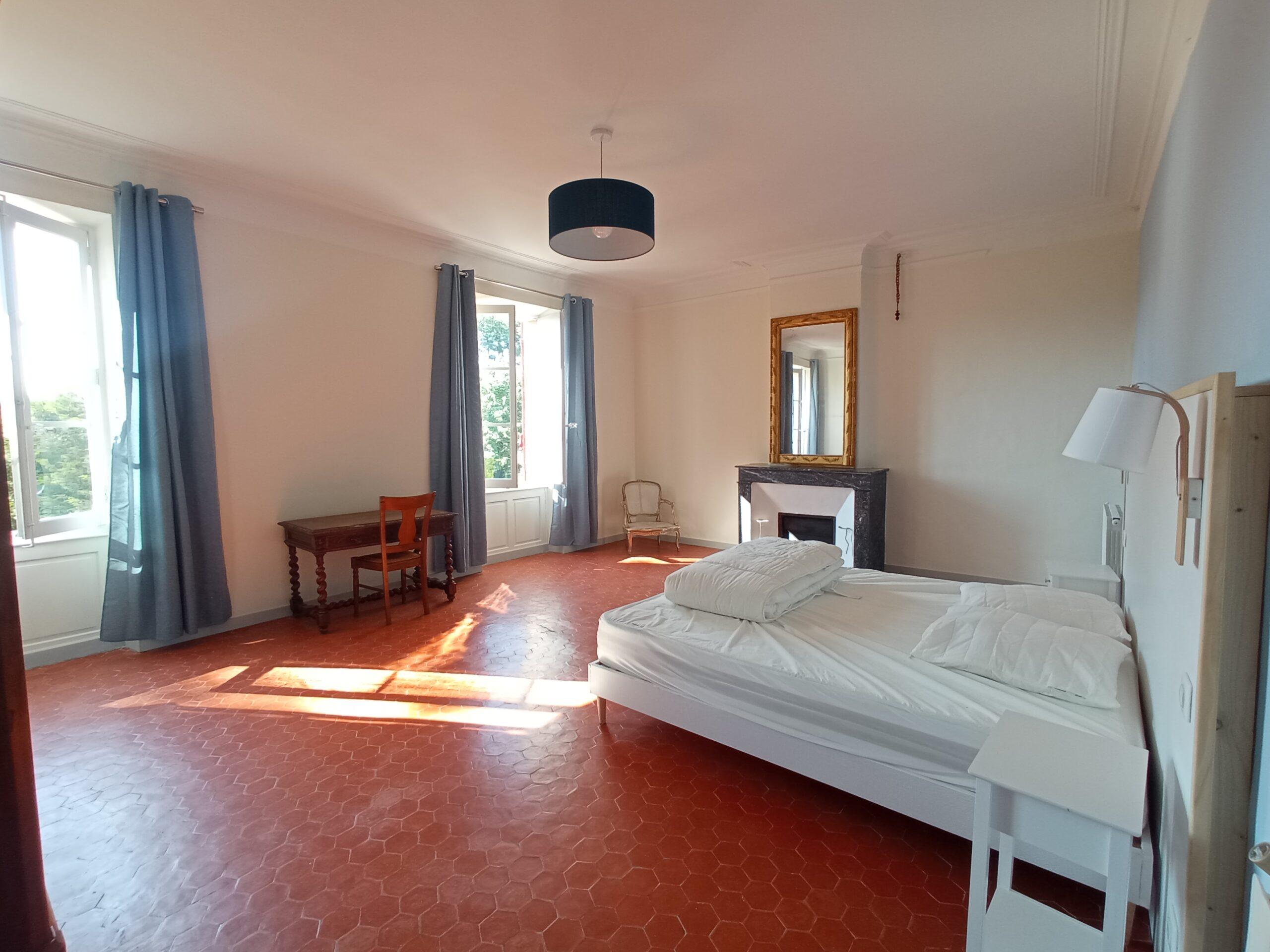 Vue intérieure de la chambre 2 dans la bastide du Domaine de Ribbe, mettant en avant un lit double, une cheminée avec miroir, un bureau ancien et un sol en tomettes.