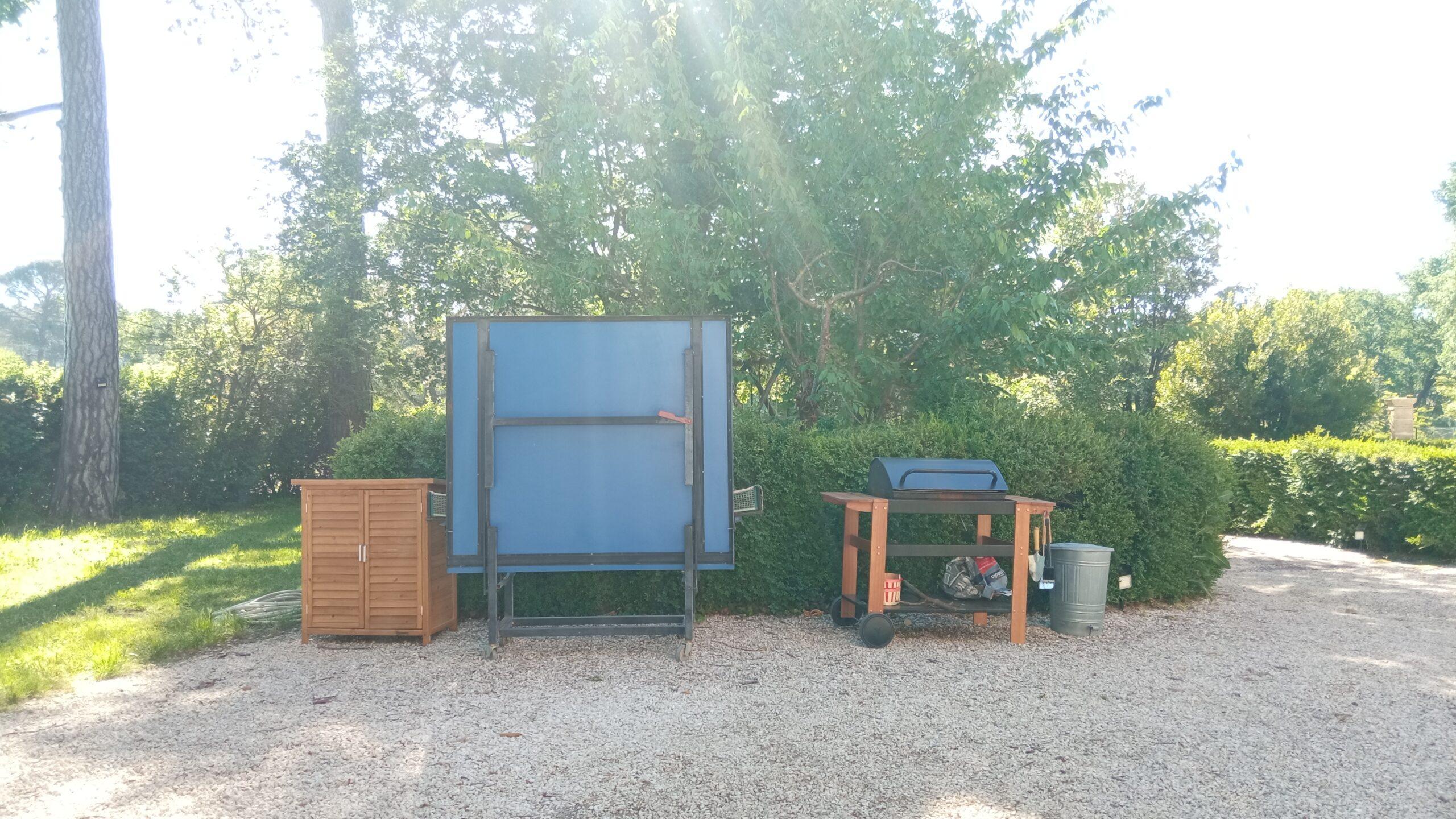 Espace de loisirs extérieur avec une table de ping-pong pliée, un barbecue et un meuble en bois sur une cour gravillonnée au Domaine de Ribbe