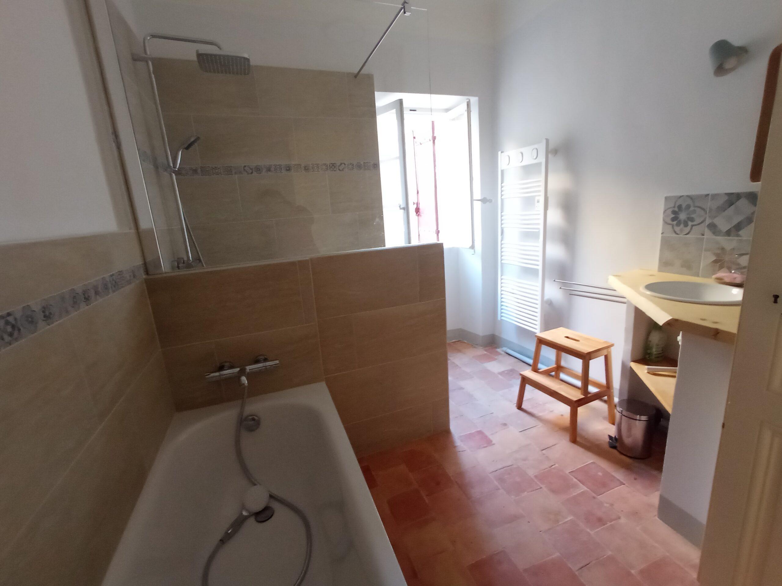 Salle de bain lumineuse pour les chambres 3 et 4 du Domaine de Ribbe, équipée d'une baignoire avec douche intégrée, d'un lavabo sur meuble en bois et d'un sol en tomettes traditionnelles.