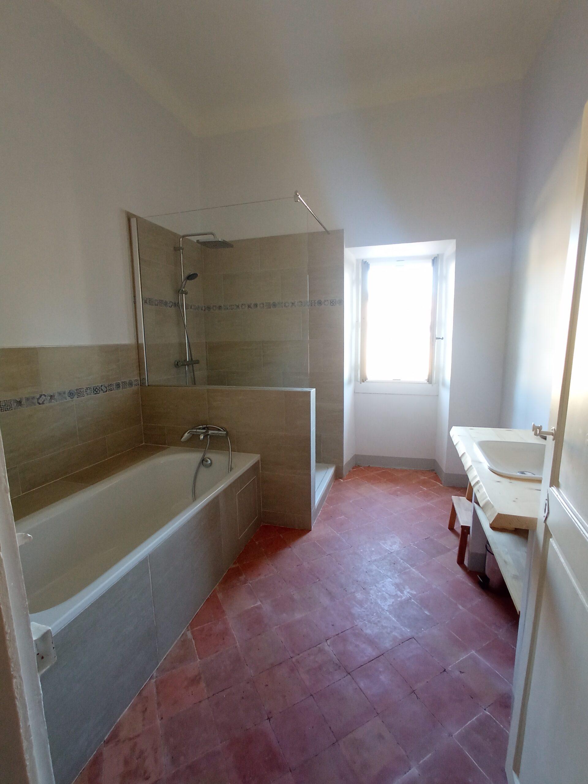 Salle de bain lumineuse desservant les chambres 7 et 8 du Domaine de Ribbe, équipée d'une baignoire, d'une douche à l'italienne et d'un lavabo sur meuble en bois, le tout sur un sol en tomettes.