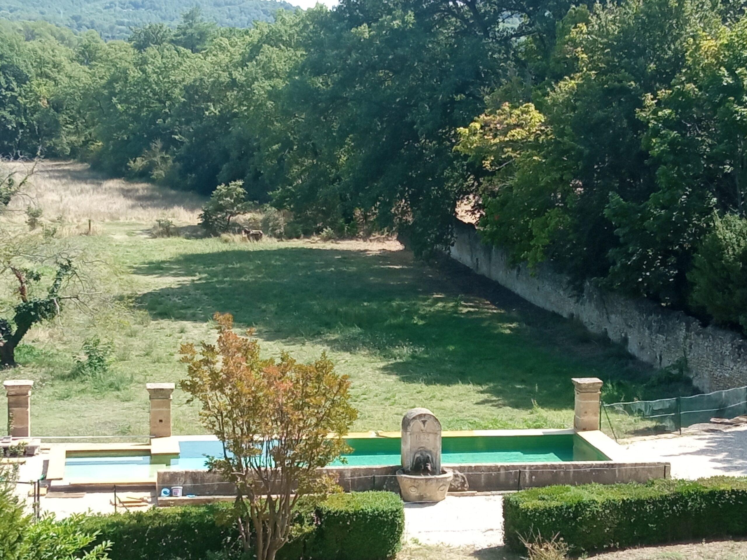 Vue en plongée du bassin de la piscine et de la fontaine historique de 1832 au Domaine de Ribbe, entourés d'arbres et de jardins.